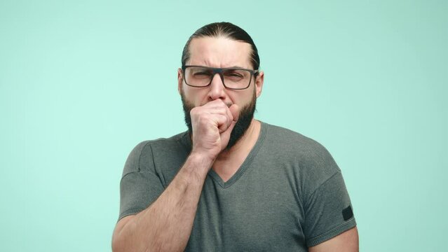 A conscientious man with glasses coughs into his fist against a teal backdrop, showing proper hygiene etiquette to prevent the spread of illness. Camera 8K RAW.