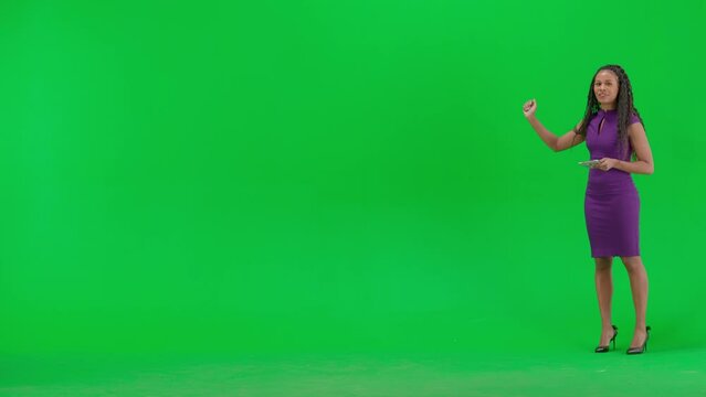 Female in dress isolated on chroma key green screen background. Full shot african american woman tv news host with tablet standing talking looking at camera.