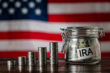 Money towers and glass jar used for saving US dollar bills and notes for IRA retirement fund on the American flag background, closeup. Finance, business, investment and money saving concept