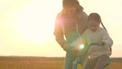 Woman teaching girl to ride bike, silhouettes at sunrise in field. Emotional support,...