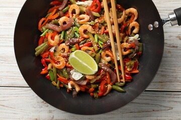 Shrimp stir fry with vegetables in wok and chopsticks on light wooden table, top view