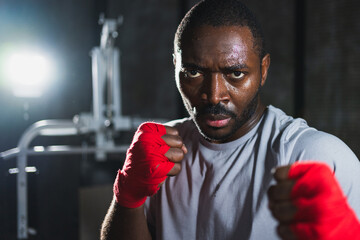 Boxer in gym. Aggressive African man fighter resting after training boxing ready for fight looking at camera. Strong sweated man training punches looking concentrated straight preparing for sparring