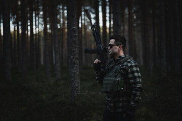 Male private military man with a modern assault rifle and body armor in the twilight forest.