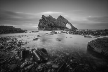 Black and white landscape of natural sea arch and stones in the foreground covered with water. Famous rock formation on the Moray Coast, Scottish Highlands, Scotland. Bow Fiddle Rock, long exposure.