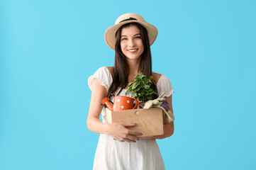 Beautiful young happy female gardener holding wooden box with tools and houseplant on blue background