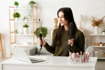 Beautiful young happy female blogger with laptop and curling iron recording video at home