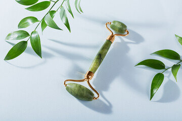 Green jade face roller and green leaves on light blue background. Massage tool for facial skin...