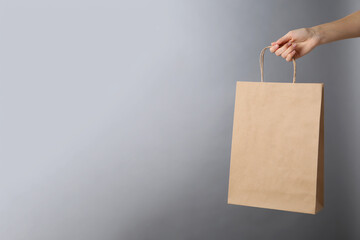 Woman holding kraft paper bag on grey background, closeup with space for text. Mockup for design