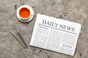 Newspaper with cup of tea and pens on grey grunge background