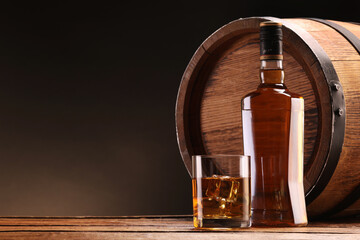 Whiskey with ice cubes in glass, bottle and barrel on wooden table against black background, space...