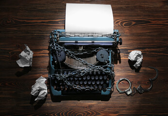 Vintage typewriter with chains, handcuffs and crumpled paper on wooden background. Printing ban...