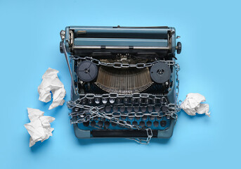 Vintage typewriter with chains and crumpled paper on blue background. Printing ban concept