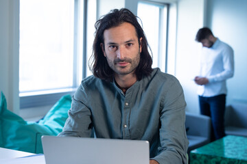 portrait d'un homme, employé de bureau ou homme d'affaire, assis au bureau devant son ordinateur portable. il regarde la caméra en souriant