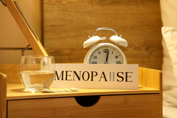 Paper with word MENOPAUSE, alarm clock, water and pills on table in bedroom at night, closeup