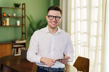 Fototapeta na wymiar A diligent man with glasses focuses on a tablet while working in a sunny, plant-filled room, epitomizing a comfortable and productive environment.