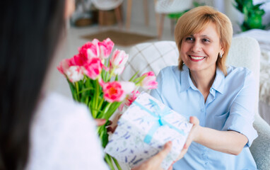 Close up photo of smiling caring grownup millennial daughter present gift and flowers to mid adult mom on womens day. Birthday, Mothers day, women's day, retired, family, relation, motherhood.