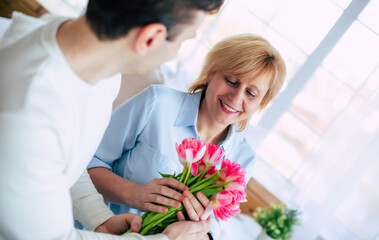 Joyful moment. Smiling mother receiving bunch of flowers from her adult son at home on the couch. Birthday, Mother's day, women day, retired, family, relation. - 773486201
