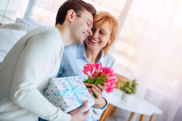 Smiling handsome adult son giving present and flowers to his mature mother at home. Birthday, Mother's day, women day, retired, family, relation. - 773486241