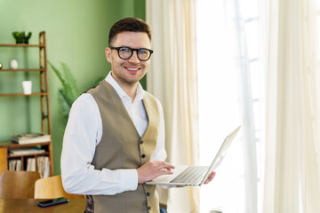 A business-savvy man in a vest and shirt holds a laptop, his confident stance signifying his...