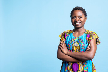 Portrait of charming person posing with arms crossed on camera, showing confidence and feeling happy with her traditional colorful outfit. African american natural woman standing in studio.