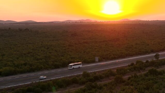 AERIAL: A lone white bus travels down the highway under a captivating crimson sunset, with a sprawling landscape around. Coach taking tourists on a road trip drives along a sunlit highway in Croatia.