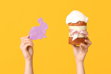 Female hands holding Easter cake and paper bunny on yellow background