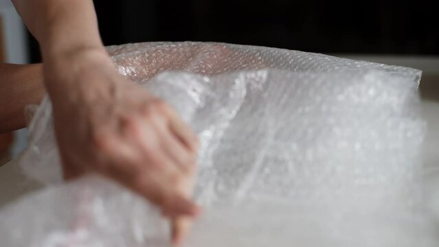 Detail cropped shot of young man pack up breakable fragile plates into wrapping bubble plastic preparing to move to new home. Closeup of pack up plates into wrapping bubble plastic, slow motion