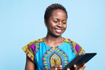African american girl is pleased to use tablet against blue backdrop, access webpages on her mobile...