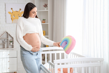 Young pregnant woman with rainbow pillow near baby crib in children's bedroom - obrazy, fototapety, plakaty