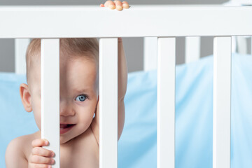 A baby peeks through the bars of a white crib