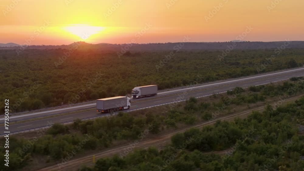 Wall mural AERIAL: A lone red bus travels down the highway under a captivating crimson sunset, with a sprawling landscape around. Coach taking tourists on a road trip drives along a sunlit highway in Croatia.