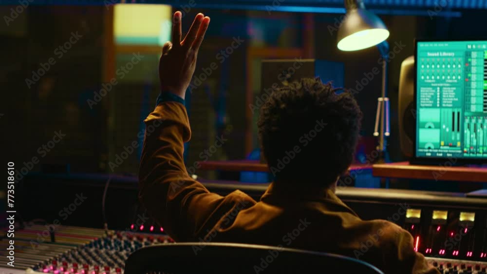 Poster Audio expert counts down for artist singing in vocal booth, recording tracks to edit them after in post production. Producer operates mixing console and buttons or sliders, control room. Camera A.