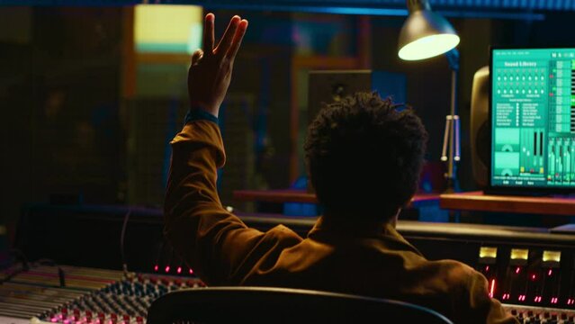 Audio expert counts down for artist singing in vocal booth, recording tracks to edit them after in post production. Producer operates mixing console and buttons or sliders, control room. Camera A.