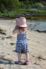 A little girl in a summer jumpsuit and a pink Panama hat walks on the seashore