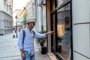 Handsome Well Dressed Young Man with Hat, Sunglasses and Backpack Standing in the City Center and Pointing at the Shopping Window