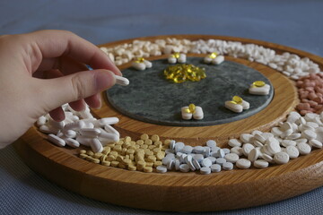 An addict's charcuterie board. One hand selecting a white pill from a selection of prescription medications arranged on a circular cheese board as if they were an appetizer for those facing addiction.