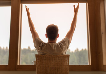 Young man with arms up looking out window in the morning feeling happy joyful and inspired  - obrazy, fototapety, plakaty
