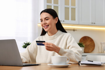 Happy young woman with credit card using laptop for shopping online at wooden table in kitchen