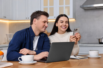 Happy couple with gadgets shopping online at wooden table in kitchen