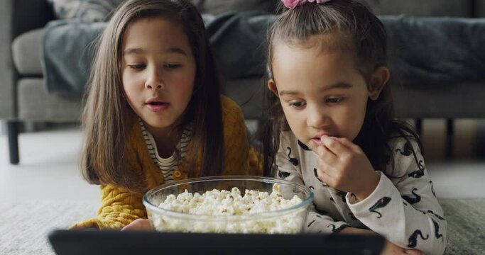 Tablet, eating popcorn and kids in family home together to watch movie, show or streaming cartoon video on app in living room. Digital technology, food snack and children on floor, girls or sisters