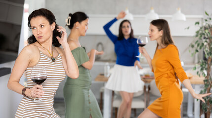 Carefree brunette in striped dress holding glass of wine and dancing with female friends at animated party in cozy home setting