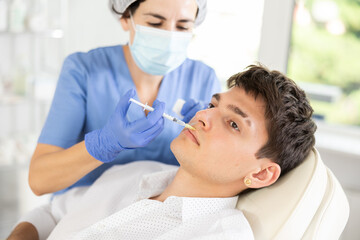 Cosmetologist in medical scrubs, cap and gloves giving filler injection to young man