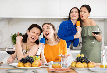 Two women joyfully sitting at set table with wine and fruits, singing into microphones at lively...