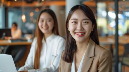 two young Asian women smiling and sitting in front of their laptops, with a modern office space as the background.