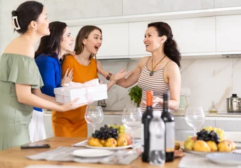 Tuinposter Girls hug and kiss on cheek when they meet, company group of three guests gives gift to birthday woman. Treats, food, alcohol and snacks on table in background. Concept of home party © JackF