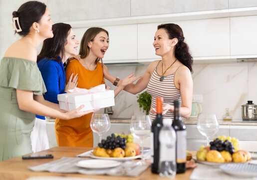 Fototapeta Girls hug and kiss on cheek when they meet, company group of three guests gives gift to birthday woman. Treats, food, alcohol and snacks on table in background. Concept of home party