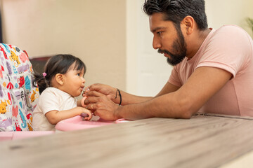 Hispanic father feeding his cheerful baby girl, Latin toddler eating in high chair, joyful moment...
