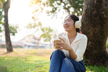 A young Asian woman wears glasses and headphones, using her mobile phone to listen to music, dance, and chat online happily, enjoying herself while relaxing on the grass in the garden. Holiday concept