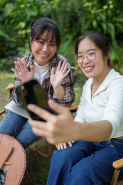 Two Asian female friends laugh and use their cell phones to chat online, say hello, take photos, and take selfies while relaxing on the grass in a green park. Holiday lifestyle concept.