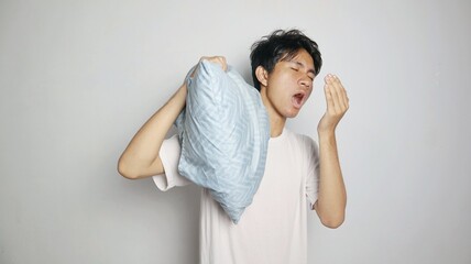 Young Asian man posing sleepy or yawning while holding a pillow behind his head, and his other hand close to his mouth